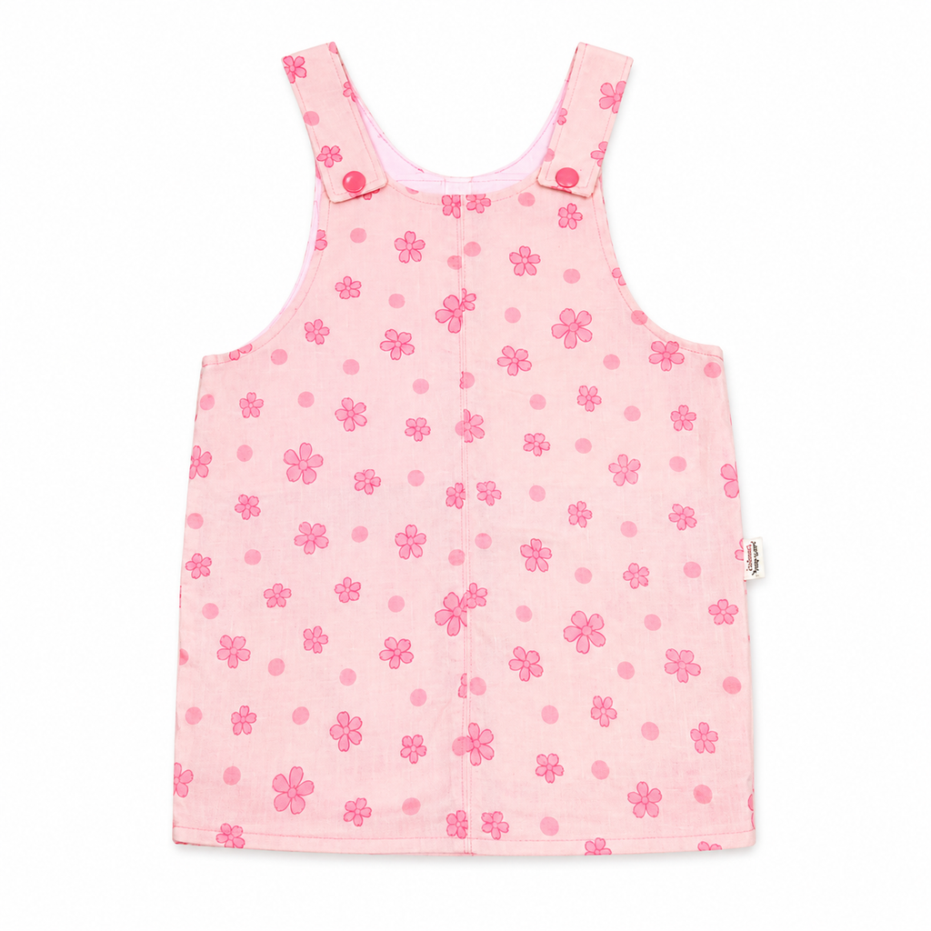 A pink sleeveless baby dress or overall-style romper with a floral and polka-dot pattern, featuring shoulder straps with snap buttons, photographed flat against a white background.