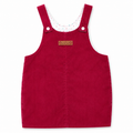 A red corduroy baby overalls dress with adjustable shoulder straps, a white inner collar with colorful polka dots, and a small brown leather brand patch on the chest.